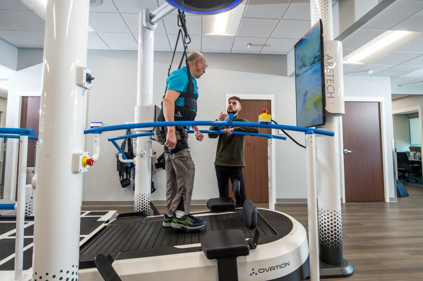 Patient in the Ovation harness pointing to the touchscreen while a therapist assists
