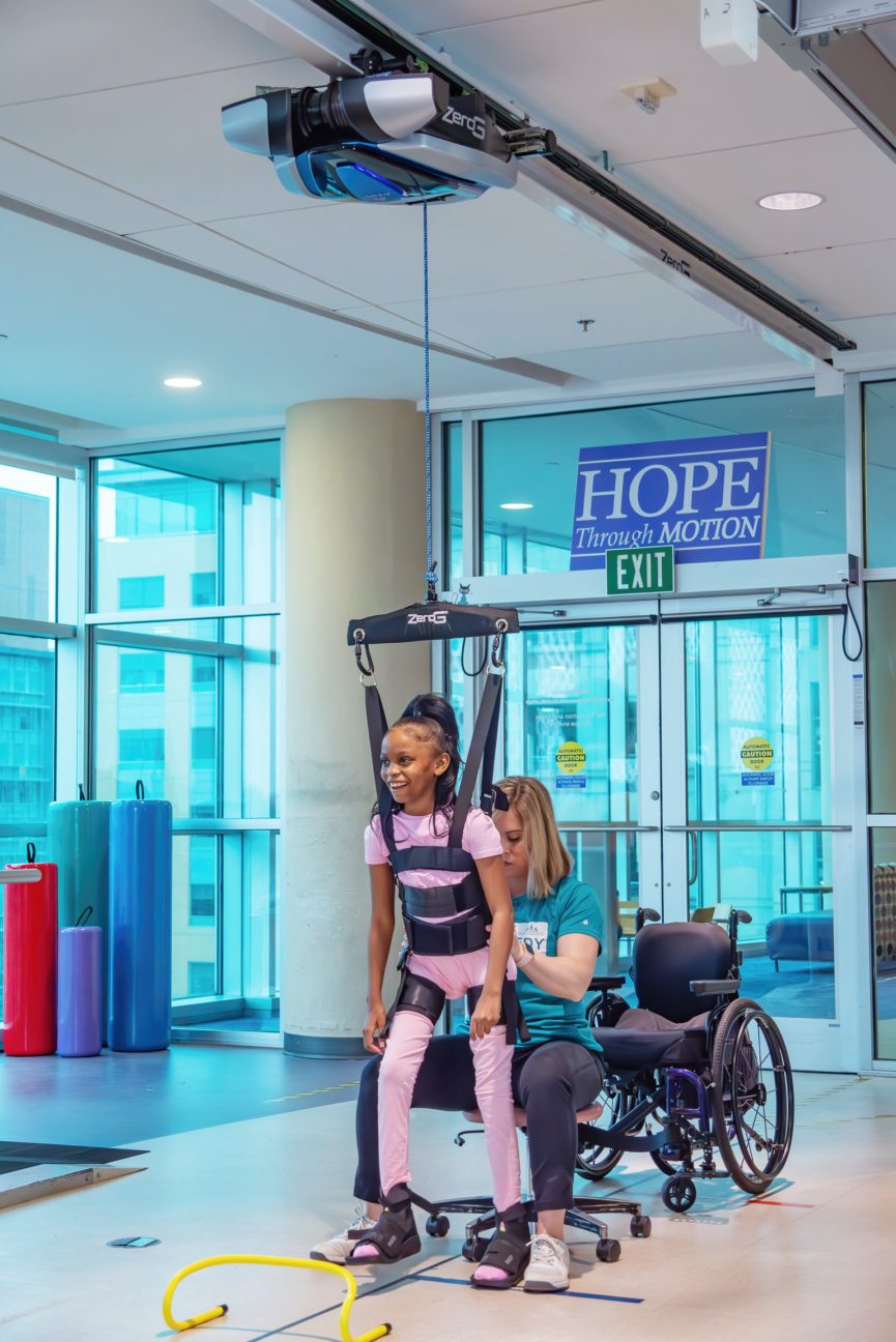 Child smiling in a ZeroG harness during a sit-to-stand exercise with a clinician