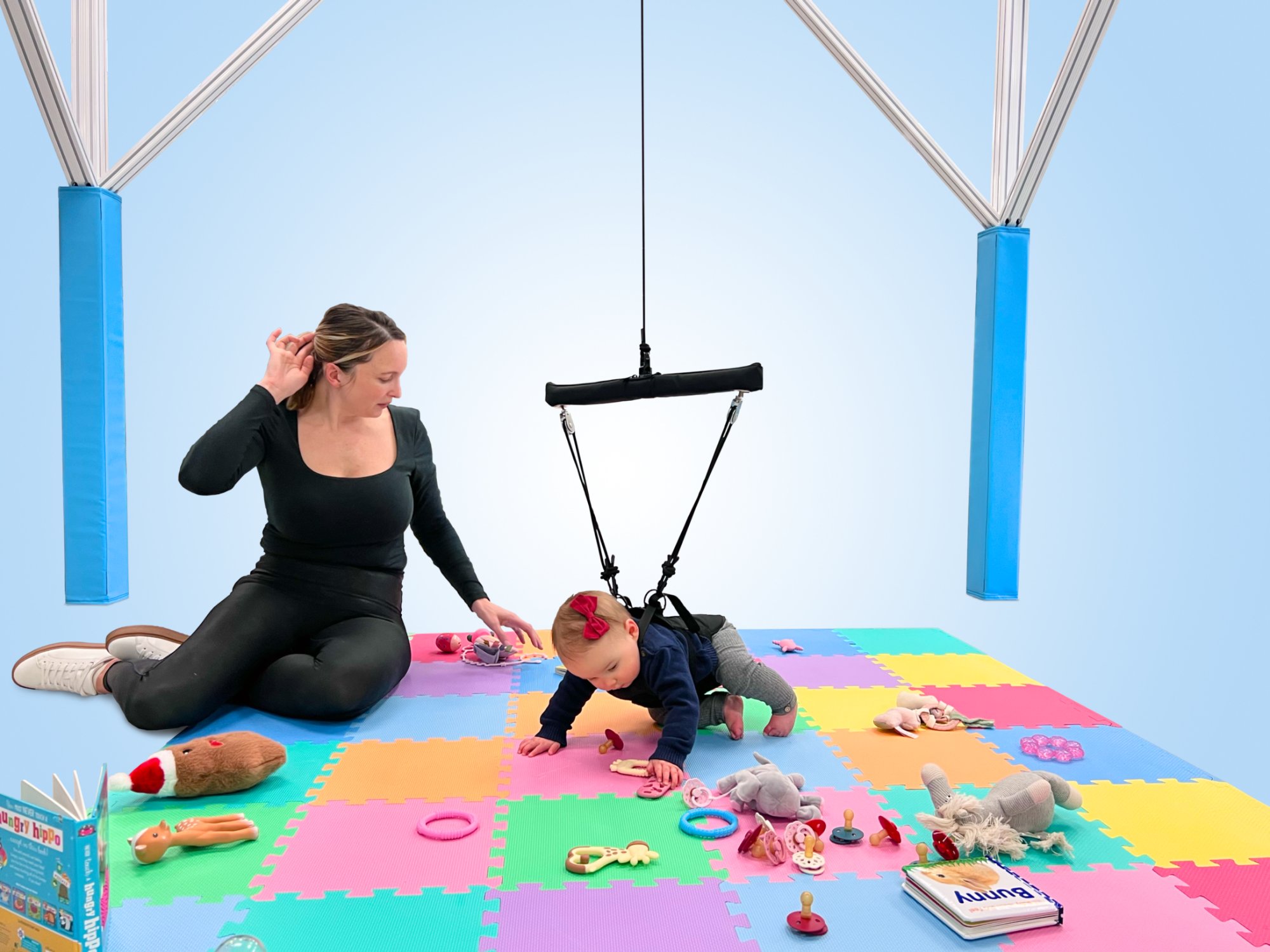 A clinician kneeling on a play mat beside an infant in a BabyG harness crawling among toys